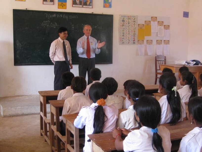 classroom with children - 2 men - one Asian and one Caucasian stood up att the front of the room talking