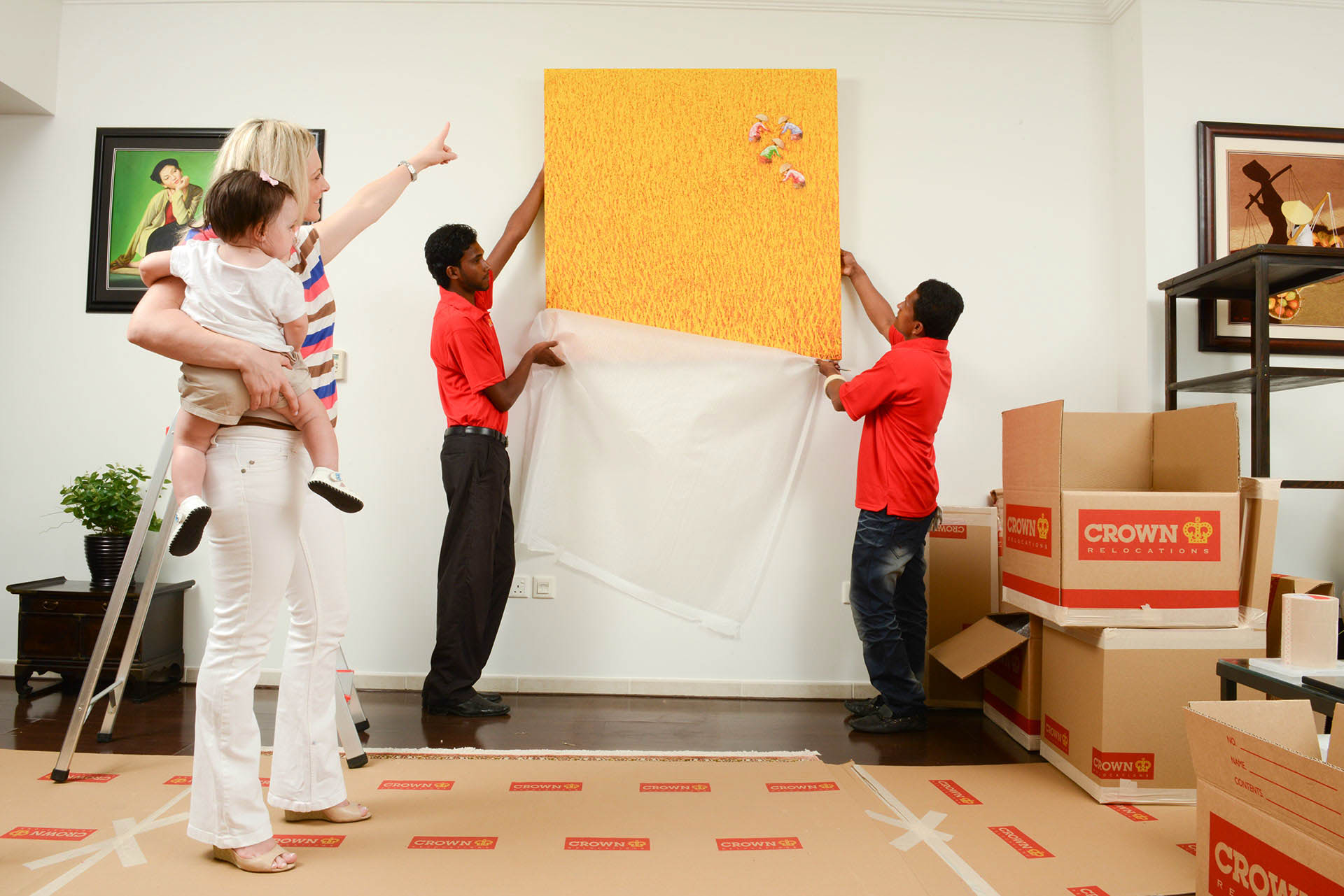packers and movers putting up a wall painting while a woman carrying a child provides direction